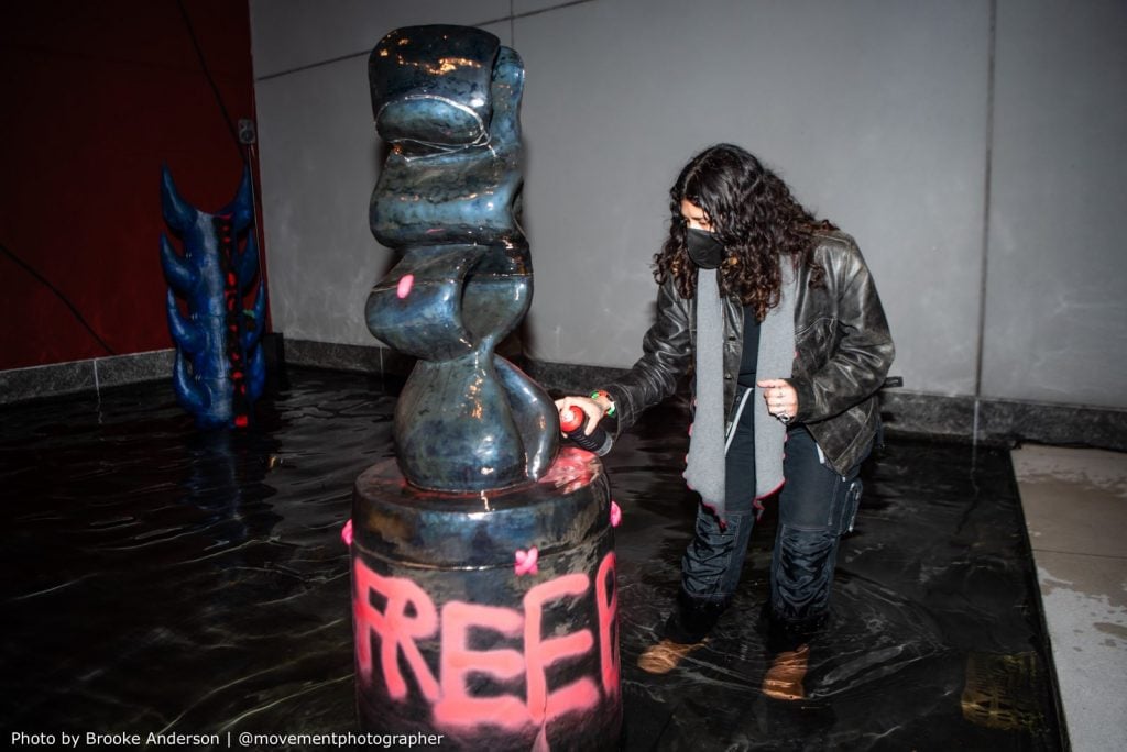 A person interacting with a sculptural fountain with a piece labeled "FREE". They are using a spray paint can on the sculpture, which is set against a glossy black base, reflecting the light and surroundings.