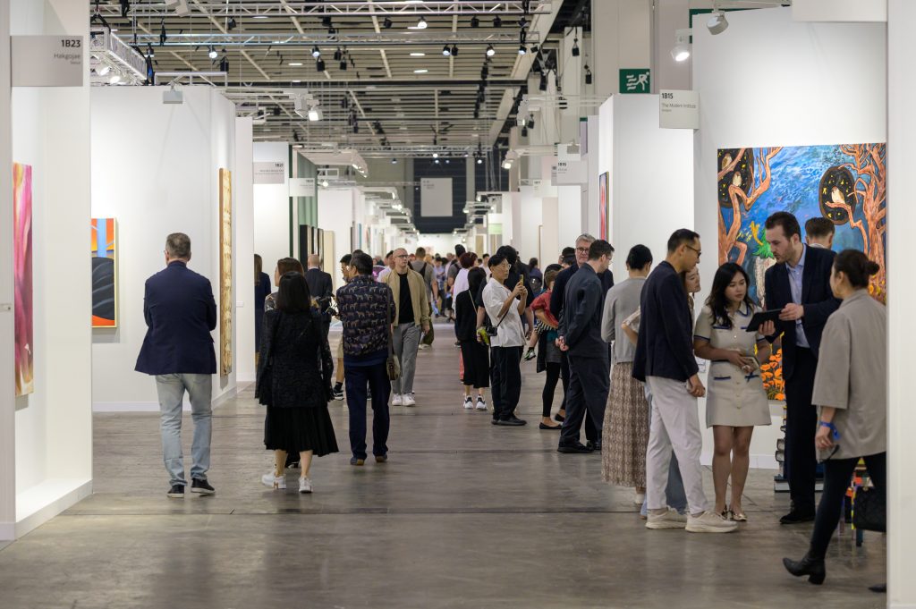 The image shows a group of people standing in an exhibition all where booths of different galleries are lined up on both sides of an aisle. The people in the photo are dressed in various styles of clothing.