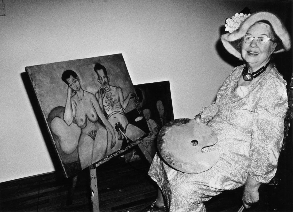 Artist Alice Neel, an elderly woman wearing a floral hat, sitting in a chair in front of a canvas she is painting