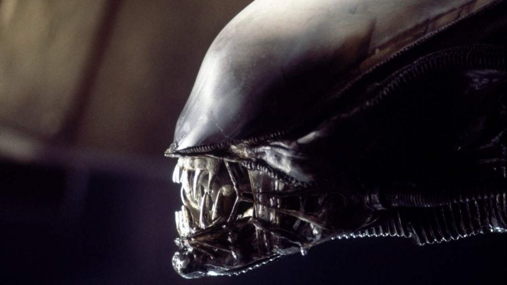 An alien with a large round head, its jaw held together by mechanical elements and its pointed teeth bared.