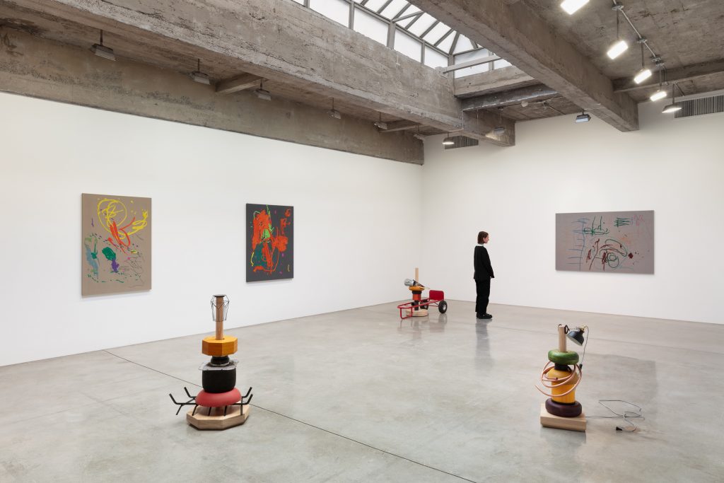 a gallery room with high ceilings. colorful abstract embroideries line the walls and three playful sculptures are on the floor. a woman in black looks at one of the embroderies.