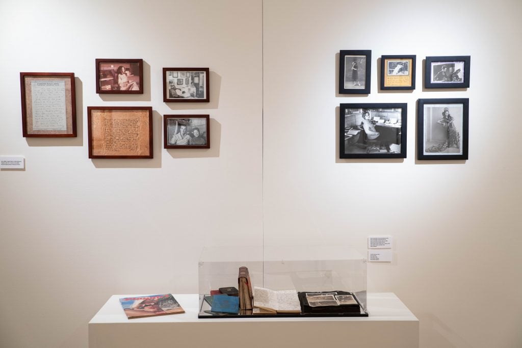 Two clusters of five pieces of rare ephemera like photos and notes mounted on a white wall behind a glass case of original diaries written by Anaïs Nin