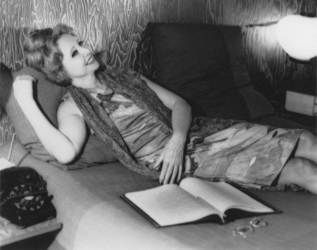 Author Anaïs Nin smiling and reclining on a bed, a file of papers open at her side.