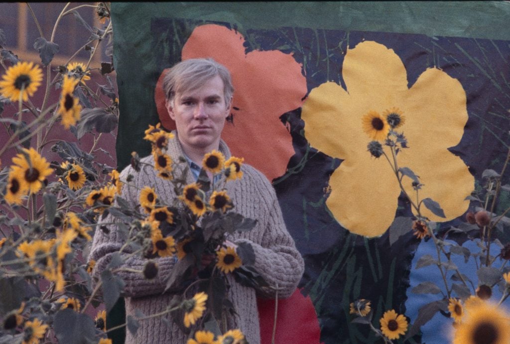 Artist Andy Warhol stands in a field of sunflowers with one of his paintings of flowers.