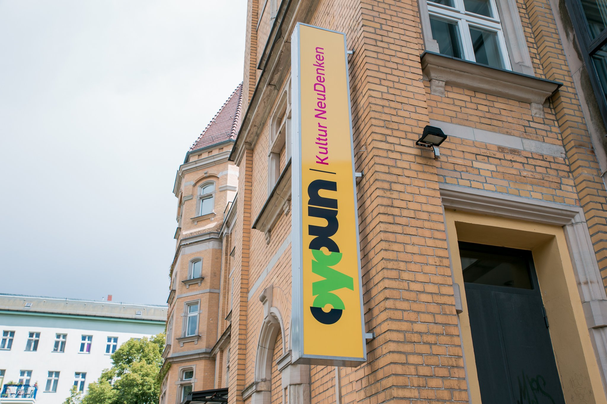 The image is of a sign on a building that reads "Oyoun Kultur NeuDenken." The building is made of light brick, located in Berlin and has windows and a clear sky in the background.