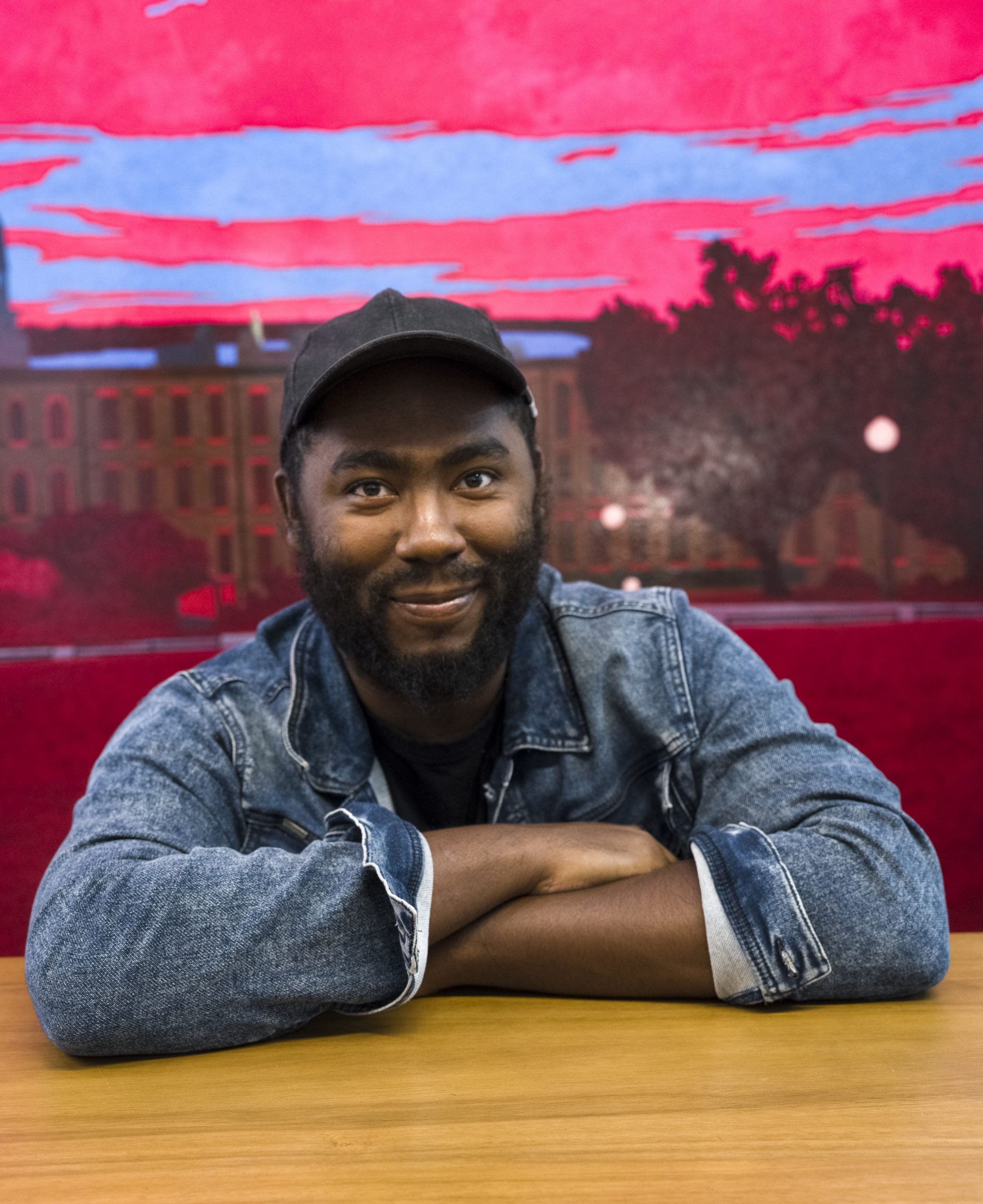 Artist Arcmanoro Niles wearing a baseball hat and jean jacket with his arms folded on a table while sitting in front of his landscape paintings with a pink and periwinkle sky and fuchsia lawn.