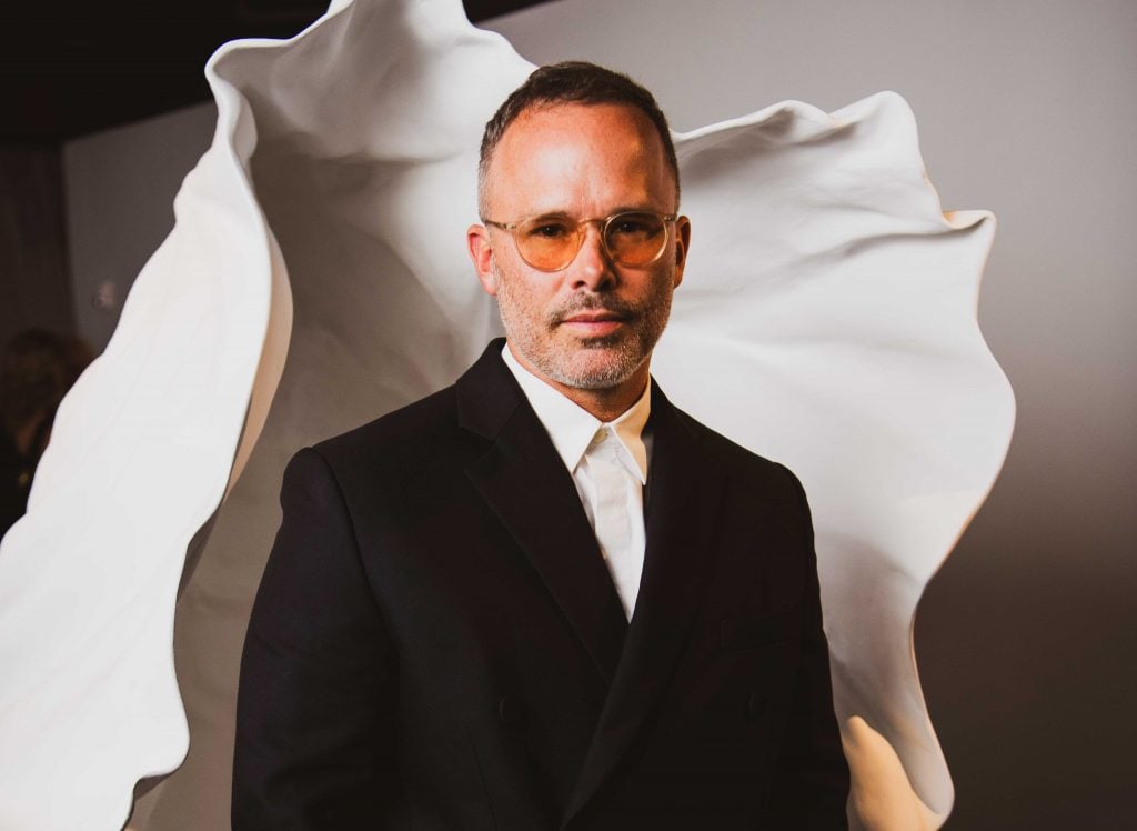 Artist Daniel Arsham, in glasses and a suit, standing in front of one of his sculptures.