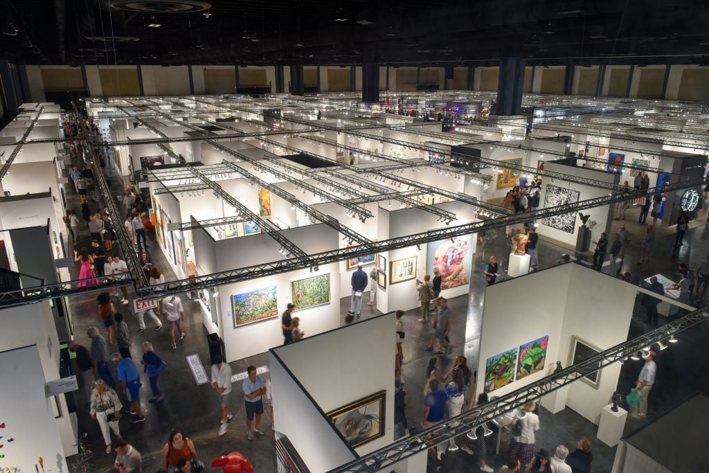 Overhead image looking across the fair floor from last year's edition, with the ceiling darkened and each booth cubicle illuminated with art.
