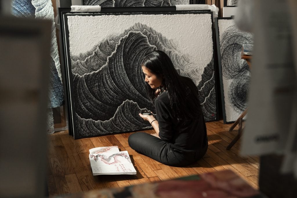 Artist Lee Hyun Joung sitting on the ground turned away working on a new artwork, and referencing a sketch. Courtesy of Galerie Sept, Brussels.