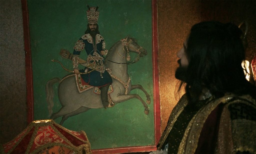 A bearded man looking at a Persian portrait of a man on horseback.