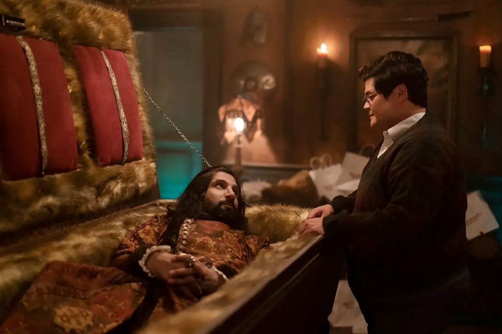 A bearded man lying down in an ornate coffin, looking up at a bespectacled man.