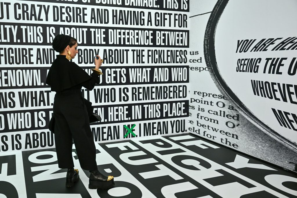 woman takes picture of Barbara Kruger text covering wall and floor of gallery