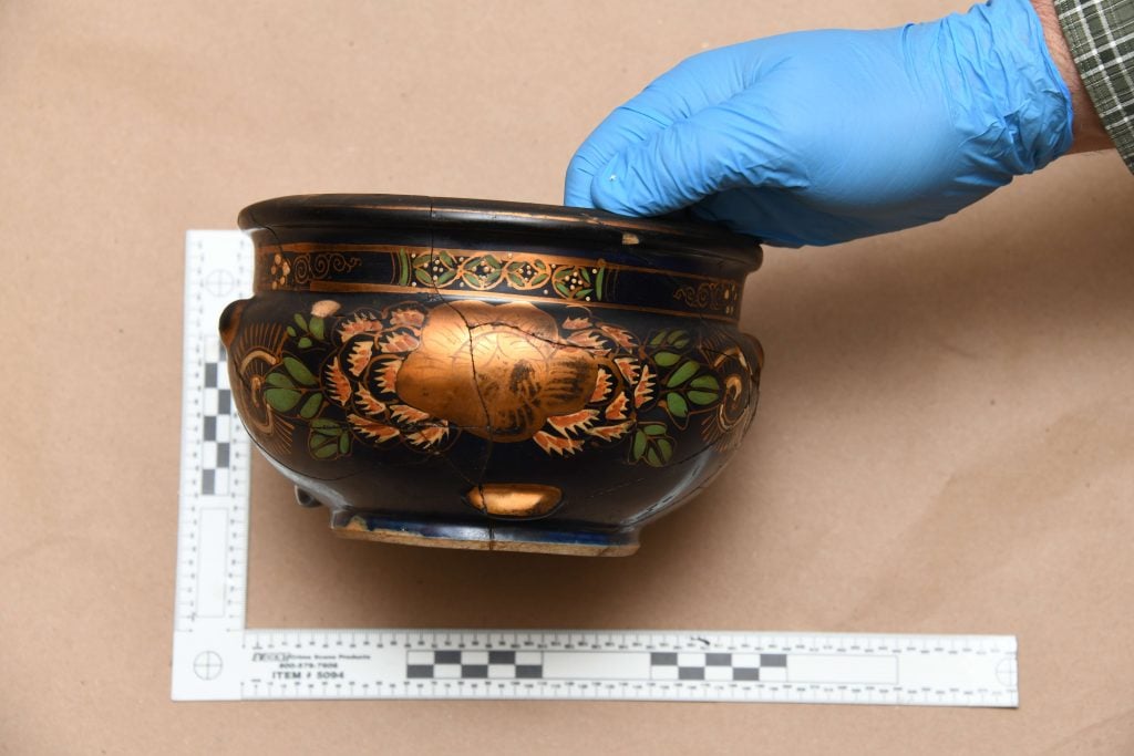 Image of a hand wearing a blue glove, holding a dark brown ceramic bowl decorated with gold and multicolored patterns, featuring a prominent flower motif. A ruler for scale is placed next to the bowl on a brown paper-covered surface.