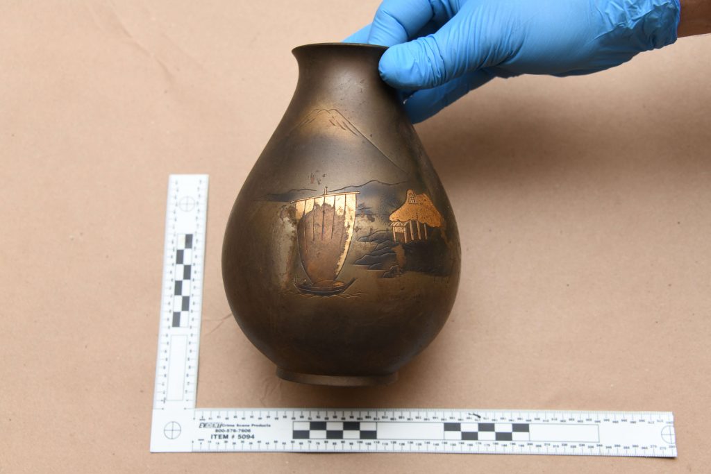 A hand in a blue glove holding a brown jug with a narrow neck and a single painted scene of a landscape with a structure and trees in gold on a black background. A ruler is placed alongside the jug for scale, on a brown paper-covered surface.