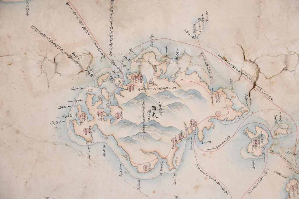 An aged map with East Asian characters, depicting a central landmass surrounded by various islands and bodies of water. Red and blue lines trace certain routes or boundaries. The paper is cracked and worn, emphasizing its antiquity.