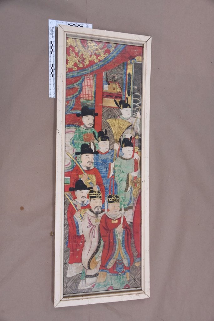 A framed, vertical scroll showing a group of East Asian figures in traditional attire, some seated and some standing, with a ruler for scale on the left edge. The painting is vibrant but shows signs of age, with a warped frame and creased paper background.