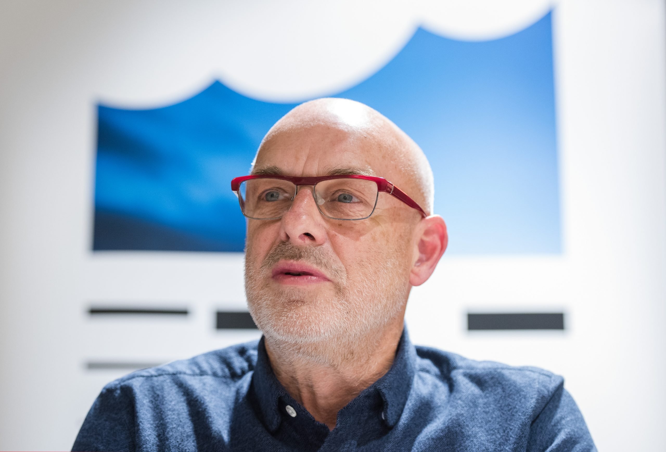 An elderly man, artist Brian Eno, pictured in close up against a backdrop of a wave-like drawing.