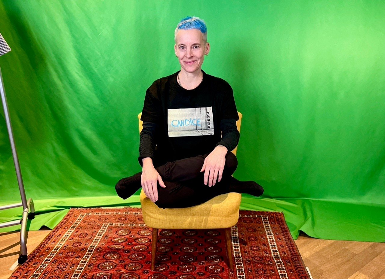 A person with short blue hair and a black long-sleeve shirt with "CANDICE" text on it is sitting cross-legged on a yellow stool. The person is on a red patterned rug over a wood floor, with a green screen background and a studio light to the left.