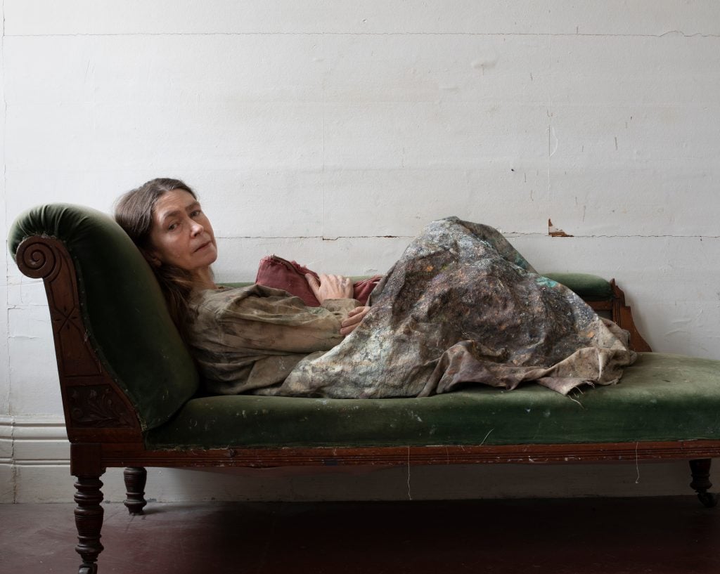 photographic portrait of celia paul laying down on a velvet forest green loveseat looking directly at the camera