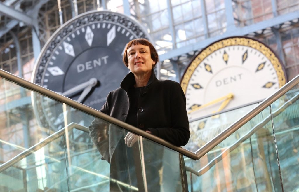 A woman posing on a balcony, an installation of clocks behind her.
