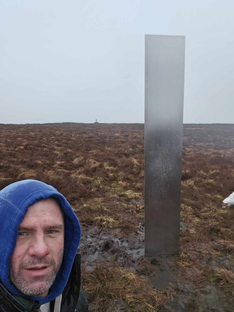 A reflective metal triangular prism, or monolith, is pictured on top of a bluff on a rainy day in Wales. A man wearing a blue hood can be seen in the bottom left corner of the frame.
