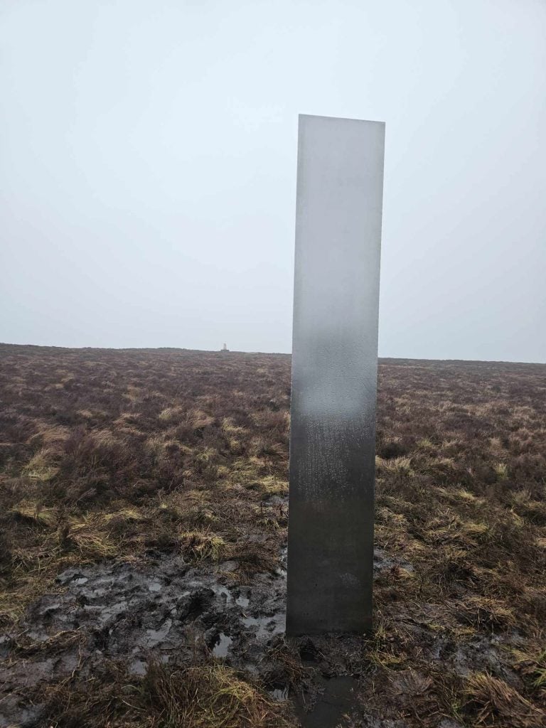 A reflective metal triangular prism, or monolith, is pictured on top of a bluff on a rainy day in Wales.
