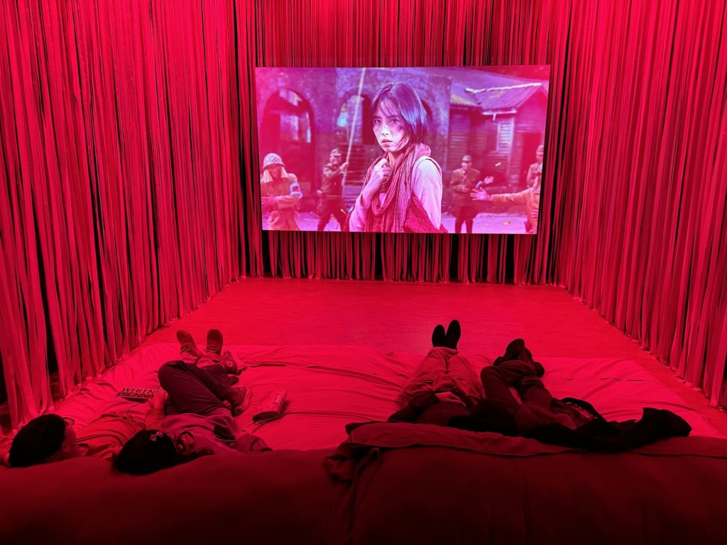 A red chamber where two people recline watching a woman onscreen