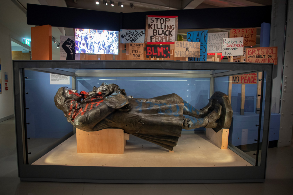 The image shows a statue of Edward Colston lying on a platform in a museum, with graffiti and messages related to Black Lives Matter and anti-racism. The statue is displayed in a museum setting.