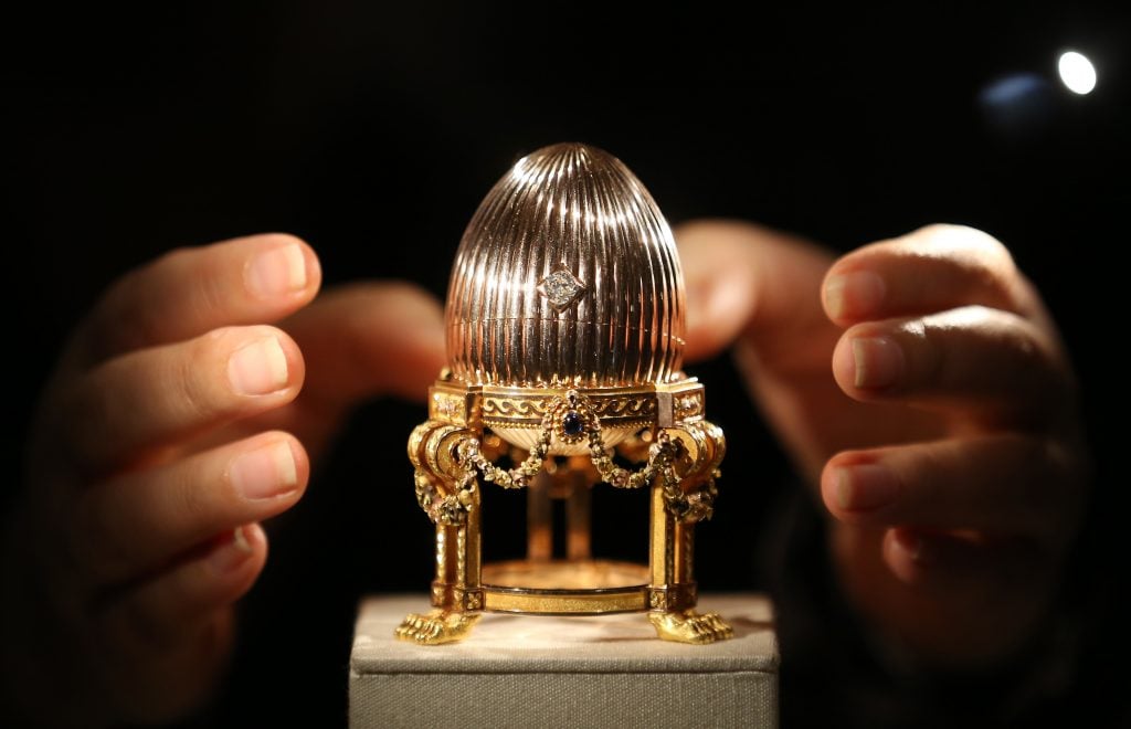 A pair of hands reaching out for an ornate gold Fabergé egg.