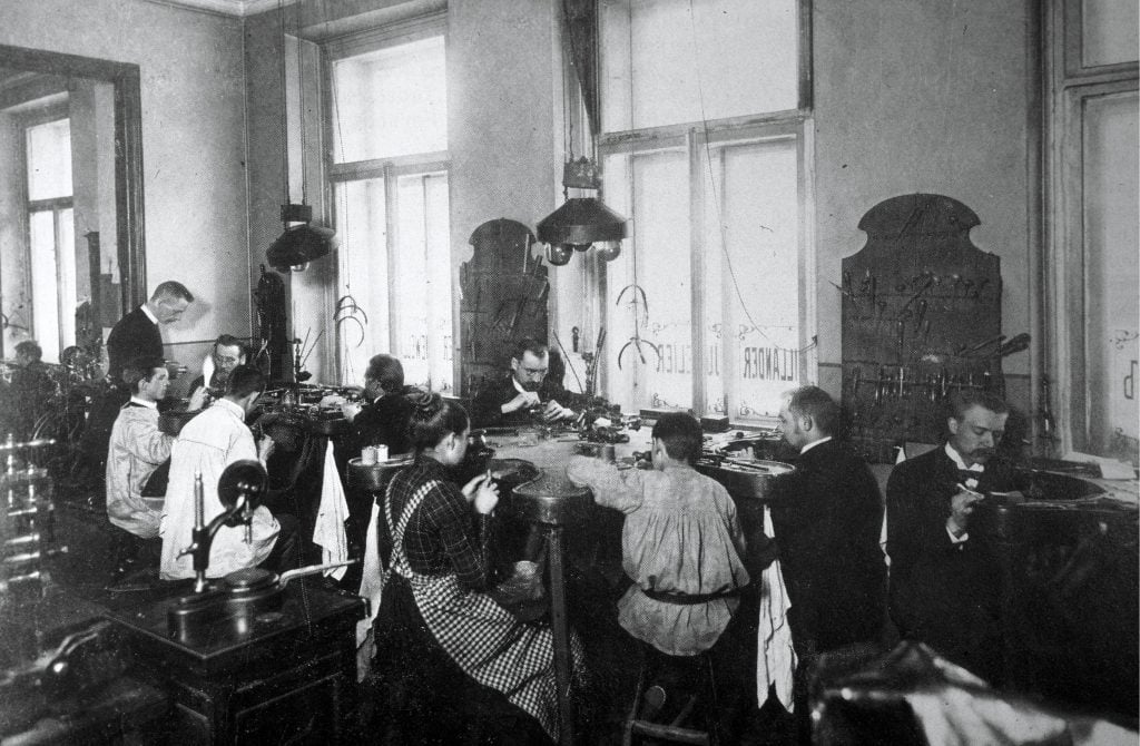 Two groups of men and women sitting around large tables at work creating Faberge eggs.