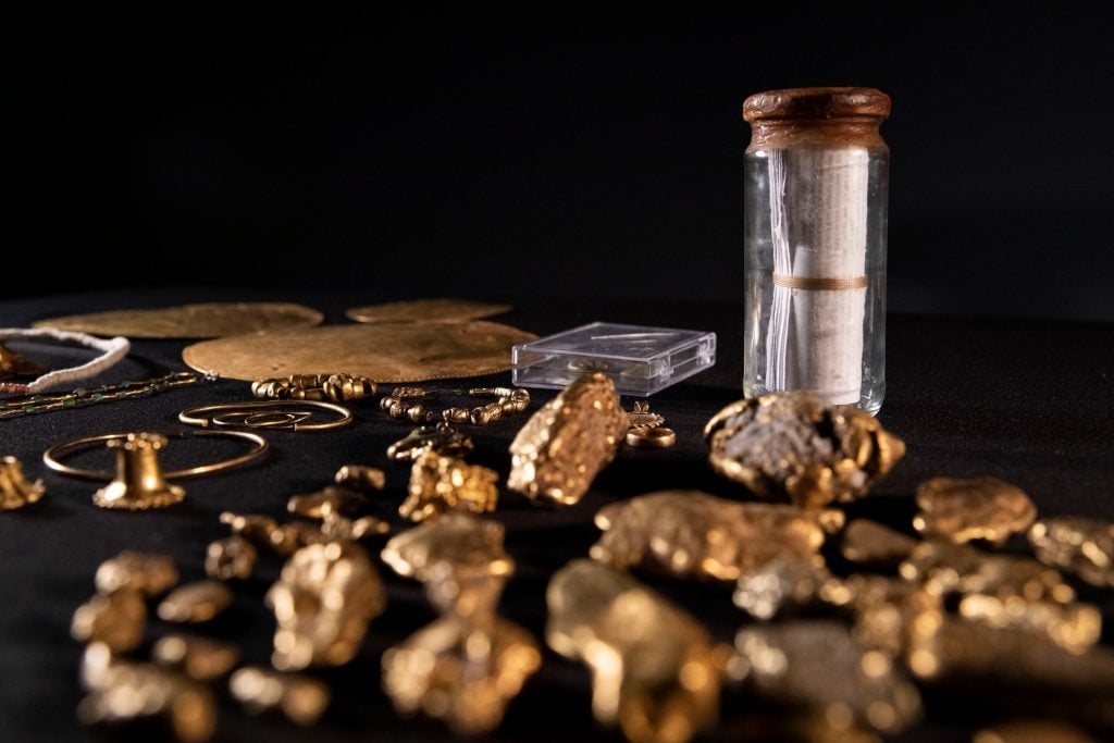 A group of gold relics and a glass jar holding a roll of paper laid out on a black surface