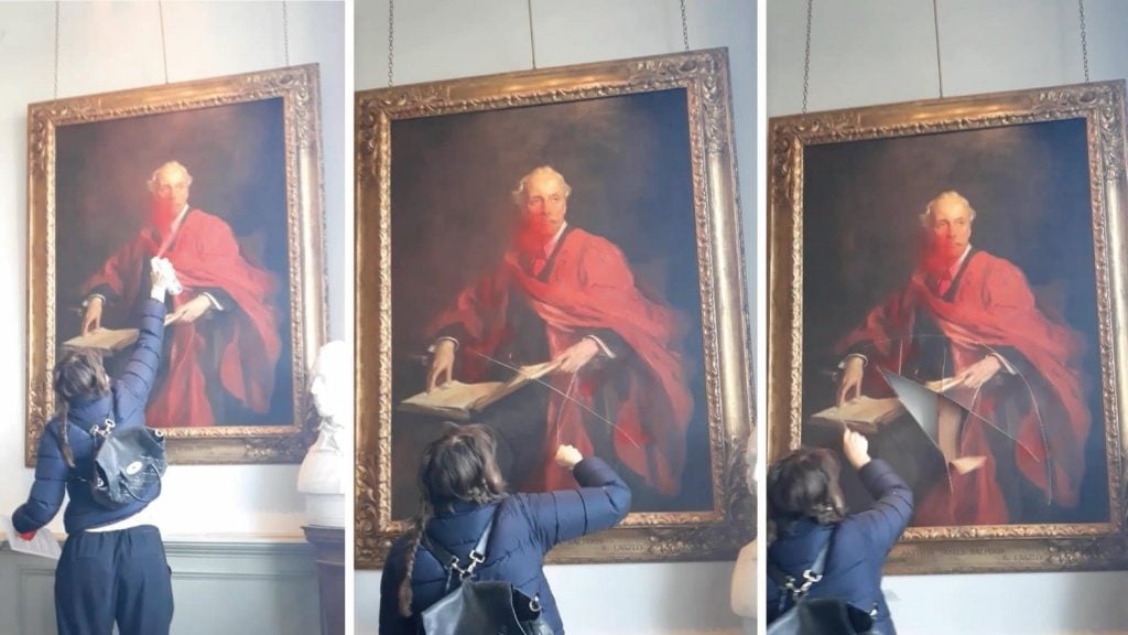 A person is seen spraypainting and slashing a painting. The painting itself is of a person dressed in red, holding what looks like a document or a book, and they appear to be looking directly out of the portrait. The artwork is in a gilded frame, and there is a plaque at the bottom, which is common for portraits that want to identify the subject or the artist, but the text is not legible in the image.