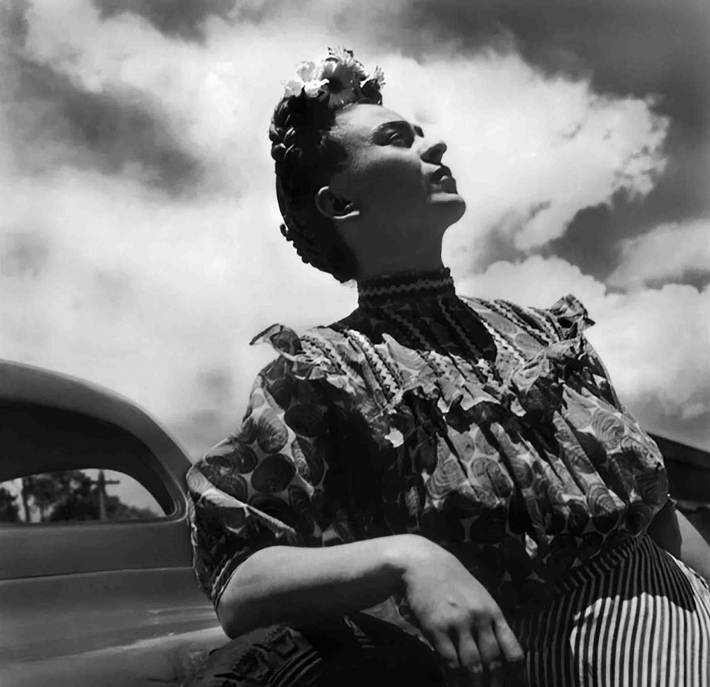 A black-and-white portrait of Mexican painter Frida Kahlo leaning against a car and gazing up at the sky.