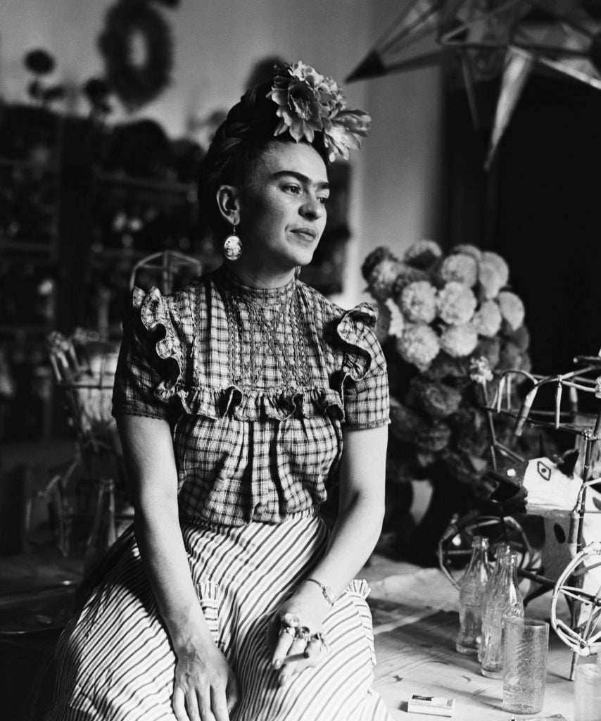 A woman, artist Frida Kahlo, in traditional Tehuana dress, leaning against a table with a vase of flowers.