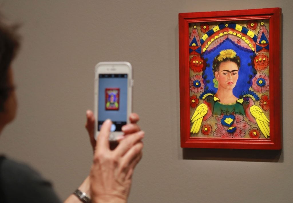 A woman holds up an iPhone camera to Frida Kahlo's colorful portrait The Frame, intricately bordered with flowers and birds, on view in a gallery.