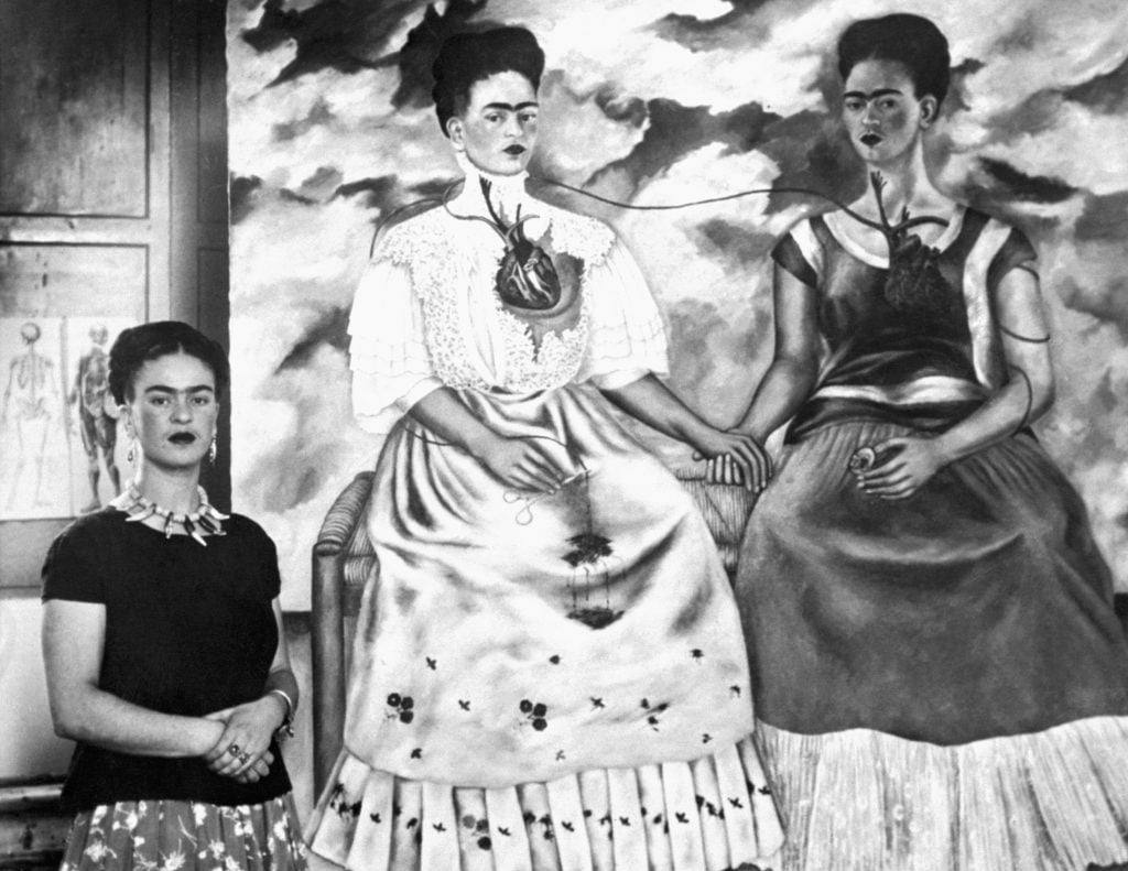 Artist Frida Kahlo standing beside her painting, The Two Fridas, depicting two versions of herself, their hearts visible through their dresses.