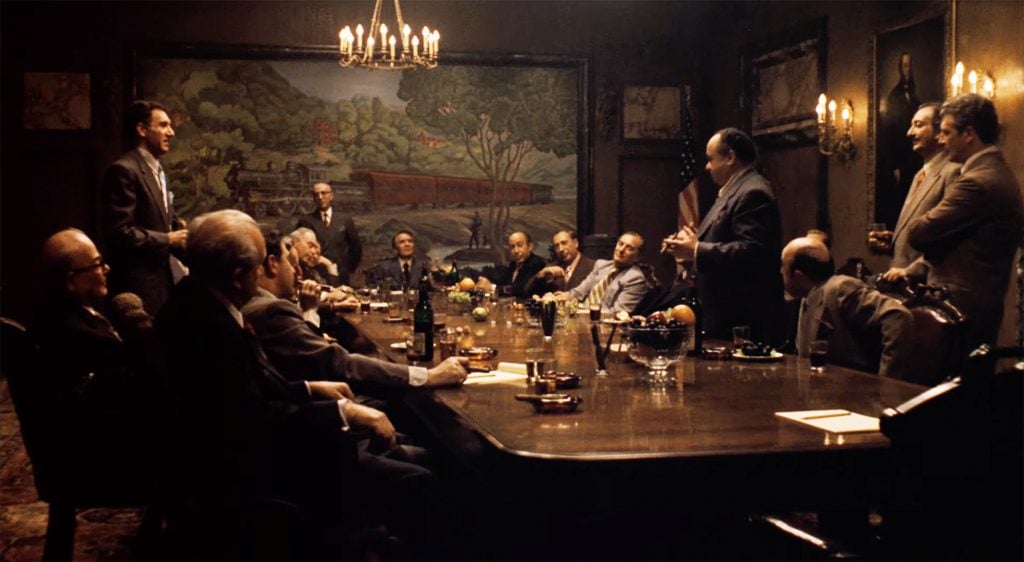 A large group of men sitting around a conference table, a large painting of a train hanging behind them.