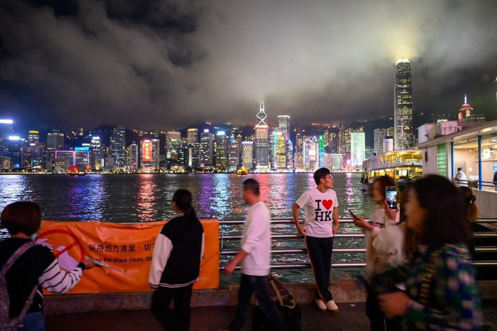 This picture shows a tourist wearing an 'I love HK' shirt poses with a view of the Hong Kong skyline in the background as seen from Kowloon on February 21, 2024 in Hong Kong, China.