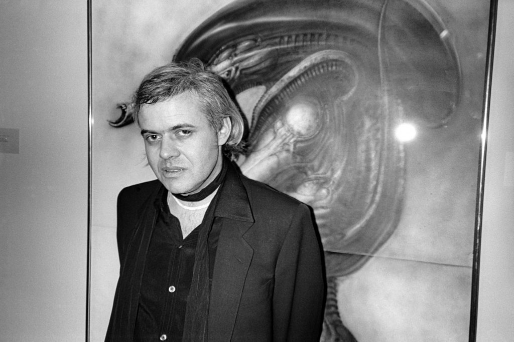 A black-and-white photo of a man, artist HR Giger, posing in front of a large drawing of a biomorphic alien.