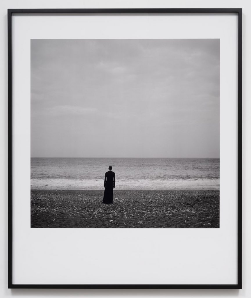 In a black-and-white photo surrounded by a white mat, a figure in dark clothing is seen on a beach. The ocean and sky are behind this person.