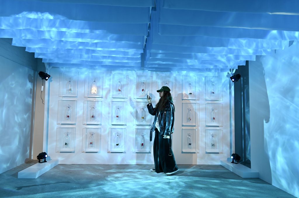 A visitor standing inside of a blue lit art installation takes a photo on their phone
