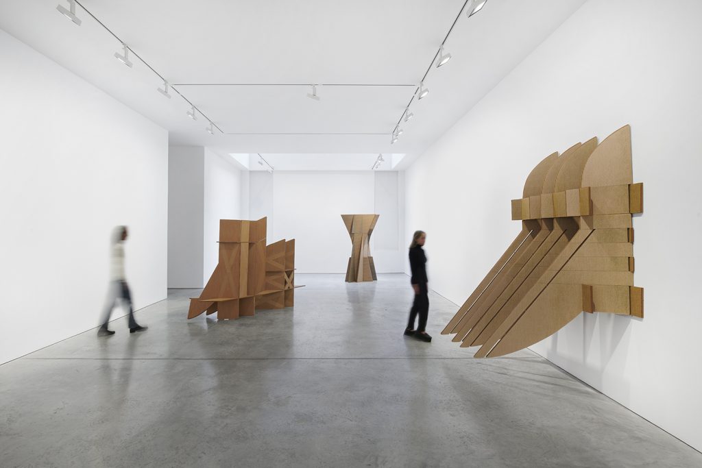 Two women walk through a gallery with large sculptures made of cardboard.
