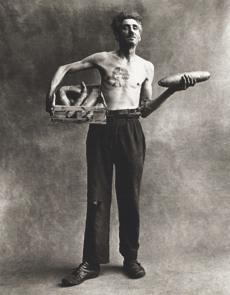 Black and white full length portrait of a shirtless man with a chest tattoo holding a basket of cucumbers under his right arm and a single cucumber in his left hand against a mottled gray seamless studio backdrop.
