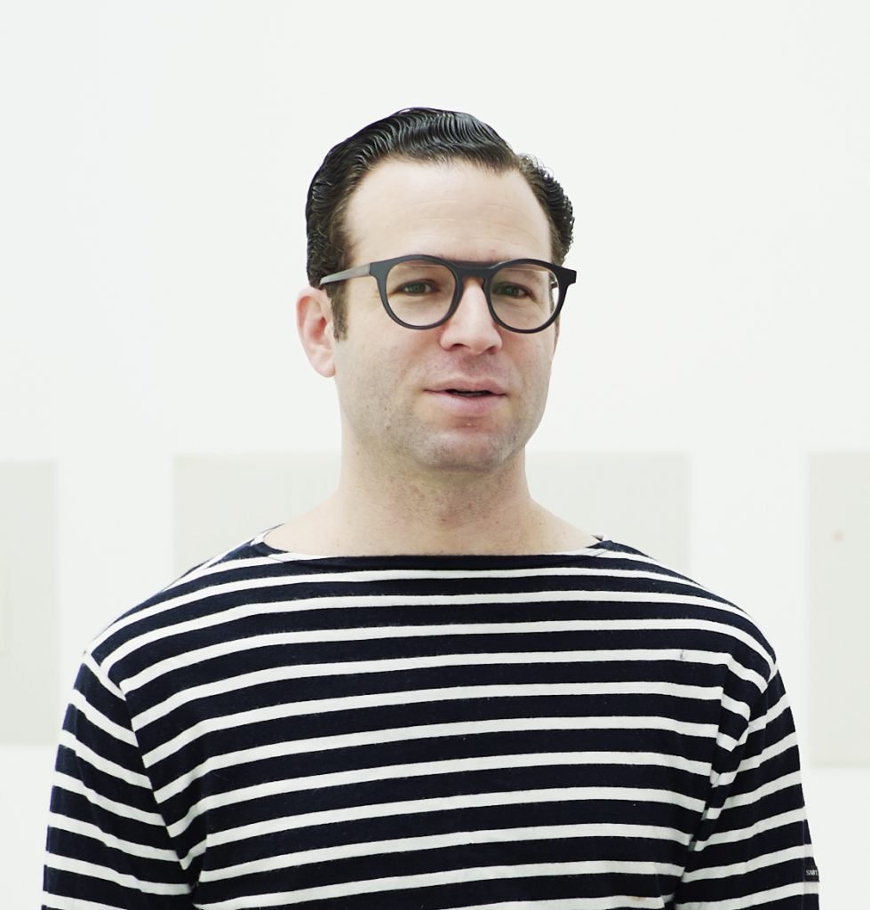 this photo shows a caucasian man wearing glasses and a black and white stripy tee shirt.