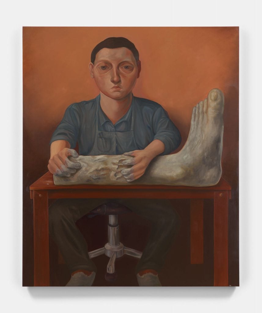 Portrait of a man sitting at a workbench that has a clay leg with foot that he is working on. His hands are working the shin of the leg, and he is sitting on a four-caster stool. The background is orange that is shadowed behind the man.