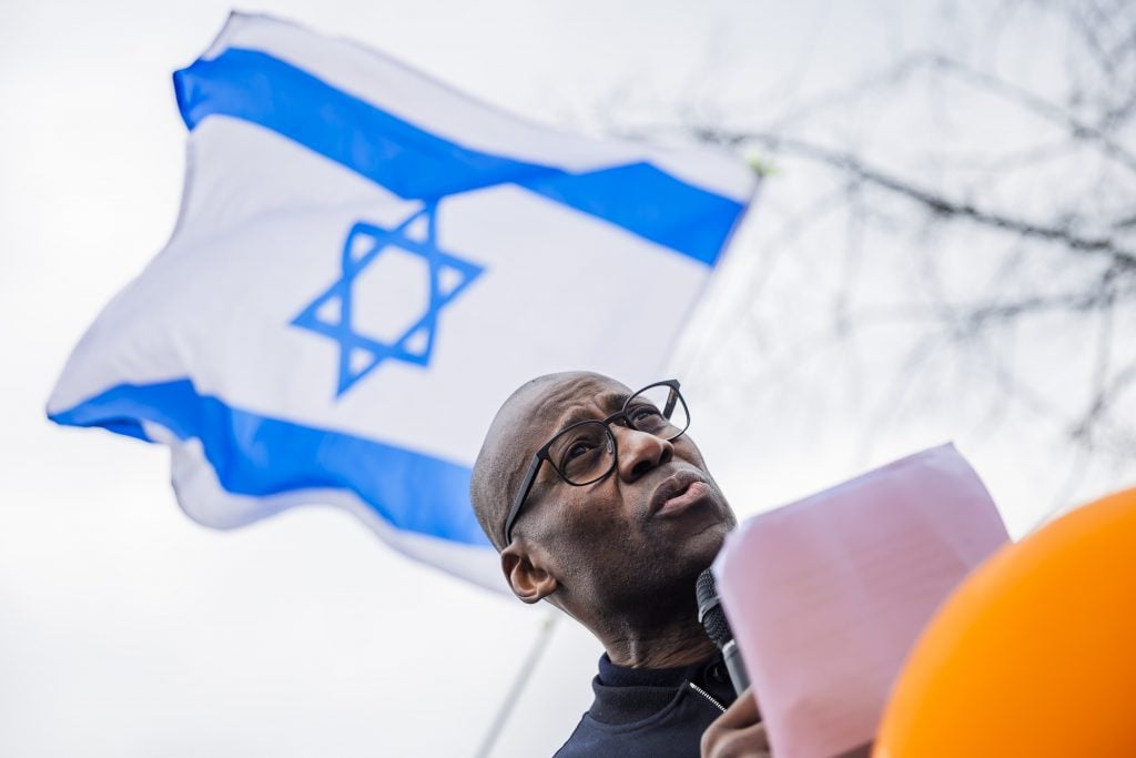 A man stands outside with a microphone in front of a waving Israel flag.