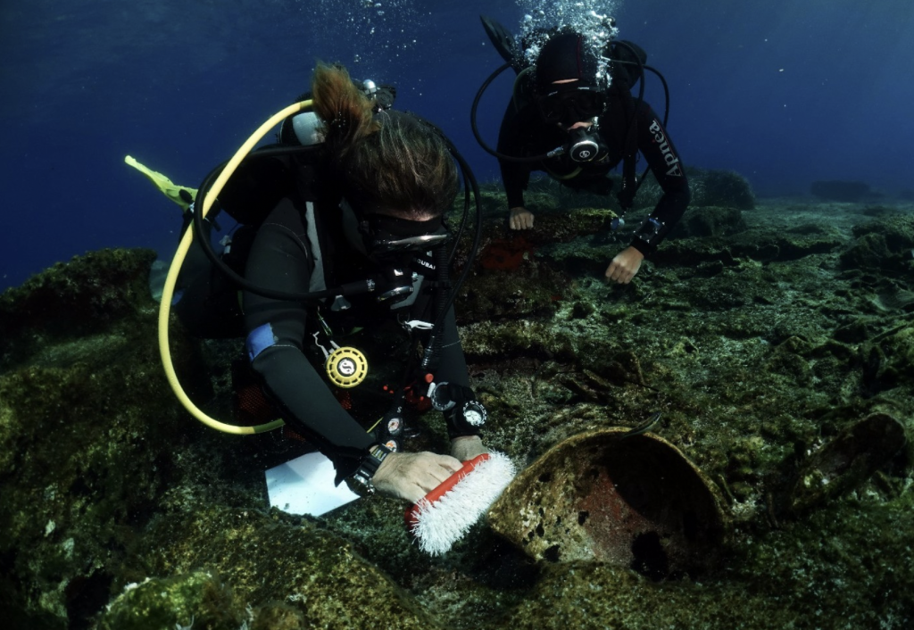 A photo of two divers examining and brushing of an ancient piece of pottery embedded in the sea floor