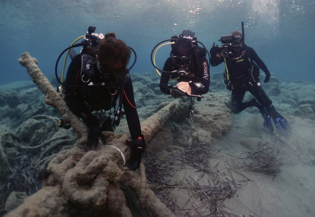 A photo of three underwater divers handling and observing the overgrown archeological remains of an Archaic period anchor