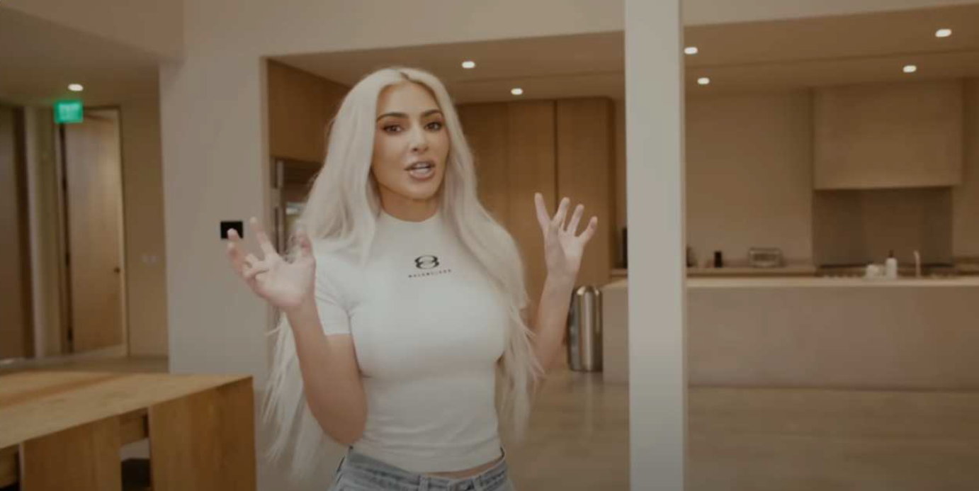 An image from a video featuring a woman speaking, with captions that read, "If you guys are furniture people." She is indoors, standing in a spacious, modern kitchen with a minimalist design. The kitchen has a large island with a wooden table extension and appears to be sparsely decorated. There's a cooking area with a backsplash and some items on the countertop. The woman is wearing a casual, form-fitting white T-shirt with a logo on the left chest area, and light-colored denim jeans. She has long, platinum blonde hair and is gesturing with her right hand, possibly emphasizing a point in her discussion. The progress bar of the video shows it's at 2 minutes and 21 seconds out of 12 minutes and 57 seconds.