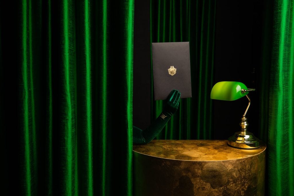 A green-gloved hand comes from behind a green curtain near a green-shaded lamp.